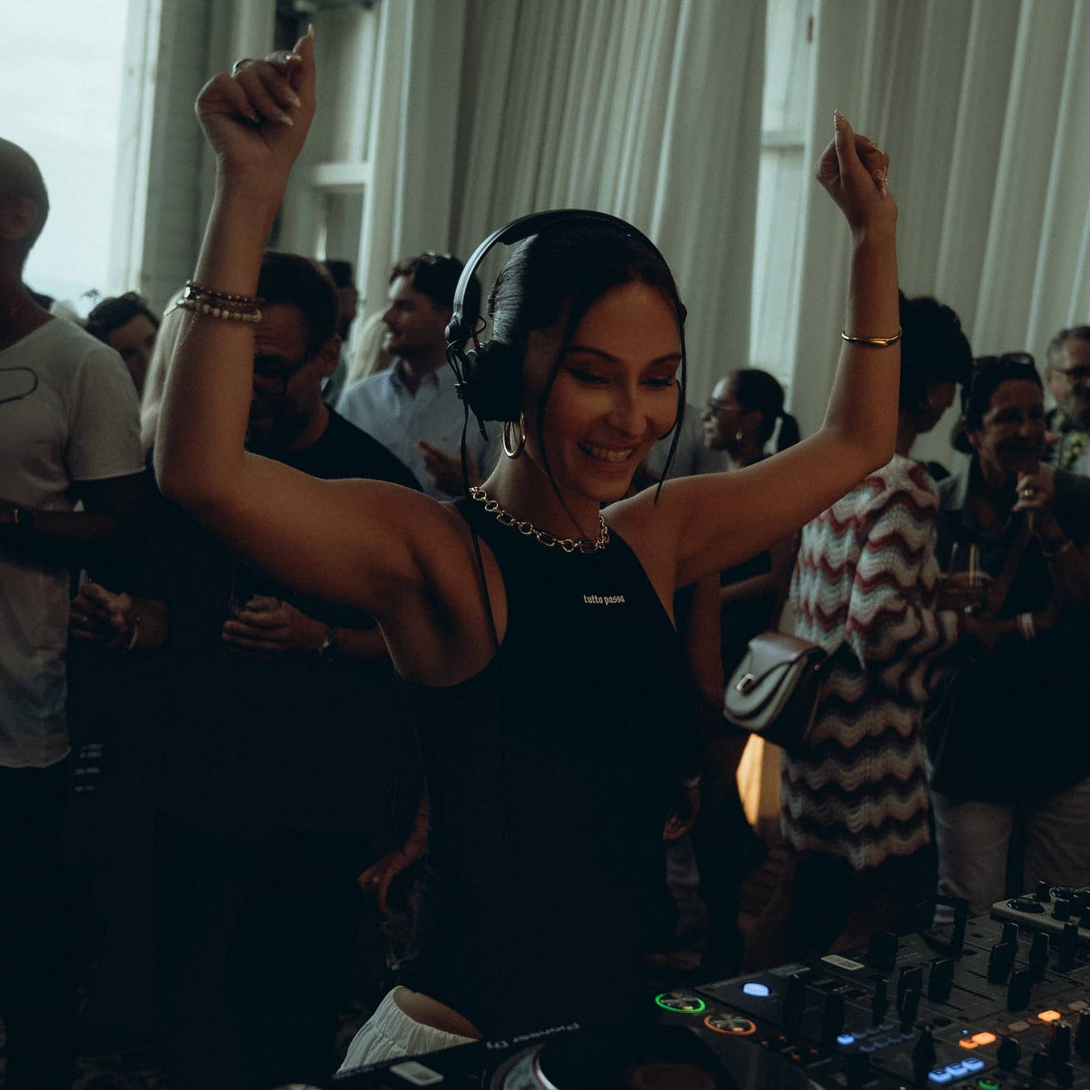 A woman DJing at a turntable with her arms raised in a crowded indoor event space, wearing a black sleeveless top and headphones.
