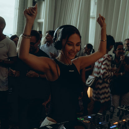 A woman DJing at a turntable with her arms raised in a crowded indoor event space, wearing a black sleeveless top and headphones.