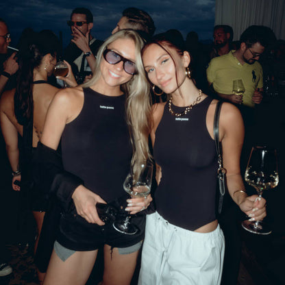 Two women posing together at a night event, wearing matching black sleeveless tops with a small white logo on the chest. The woman on the left wears black shorts and sunglasses, while the woman on the right wears white pants and a chunky gold necklace.